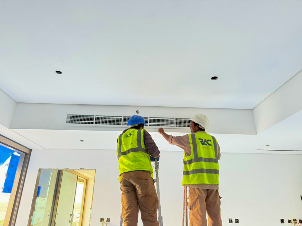 Beach Walk Iii By Imtiaz Two construction workers in safety vests install ceiling fixtures, standing on a ladder in a newly built room.